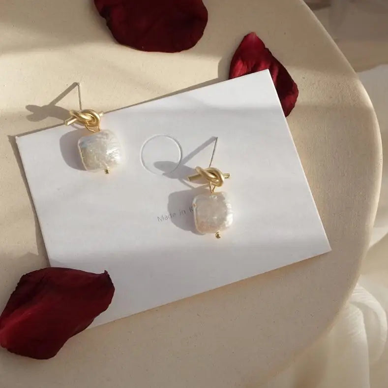 Bound in Grace Earrings displayed on a white card with soft shadows and rose petals, emphasizing their delicate pearl and gold design.