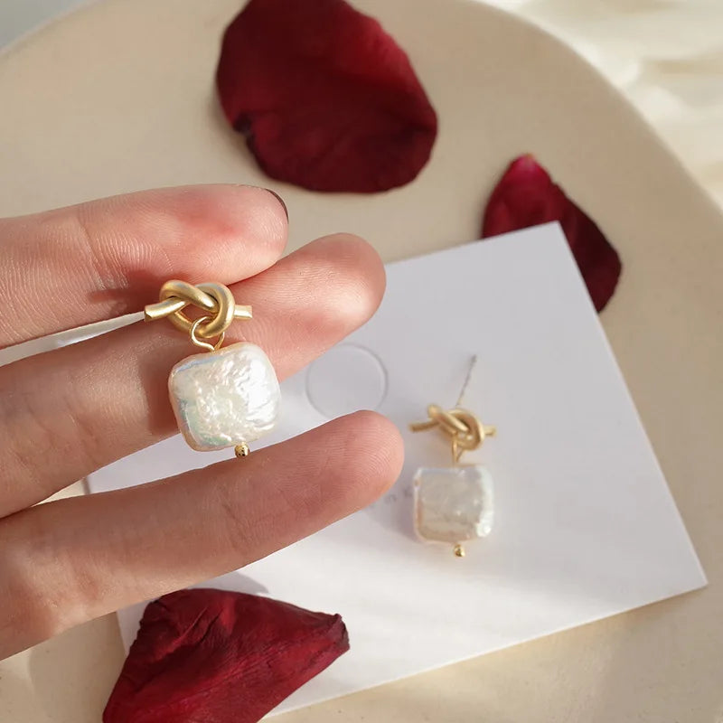 Close-up of Bound in Grace Earrings with gold knot and square pearl drop, placed on a white card with red rose petals.