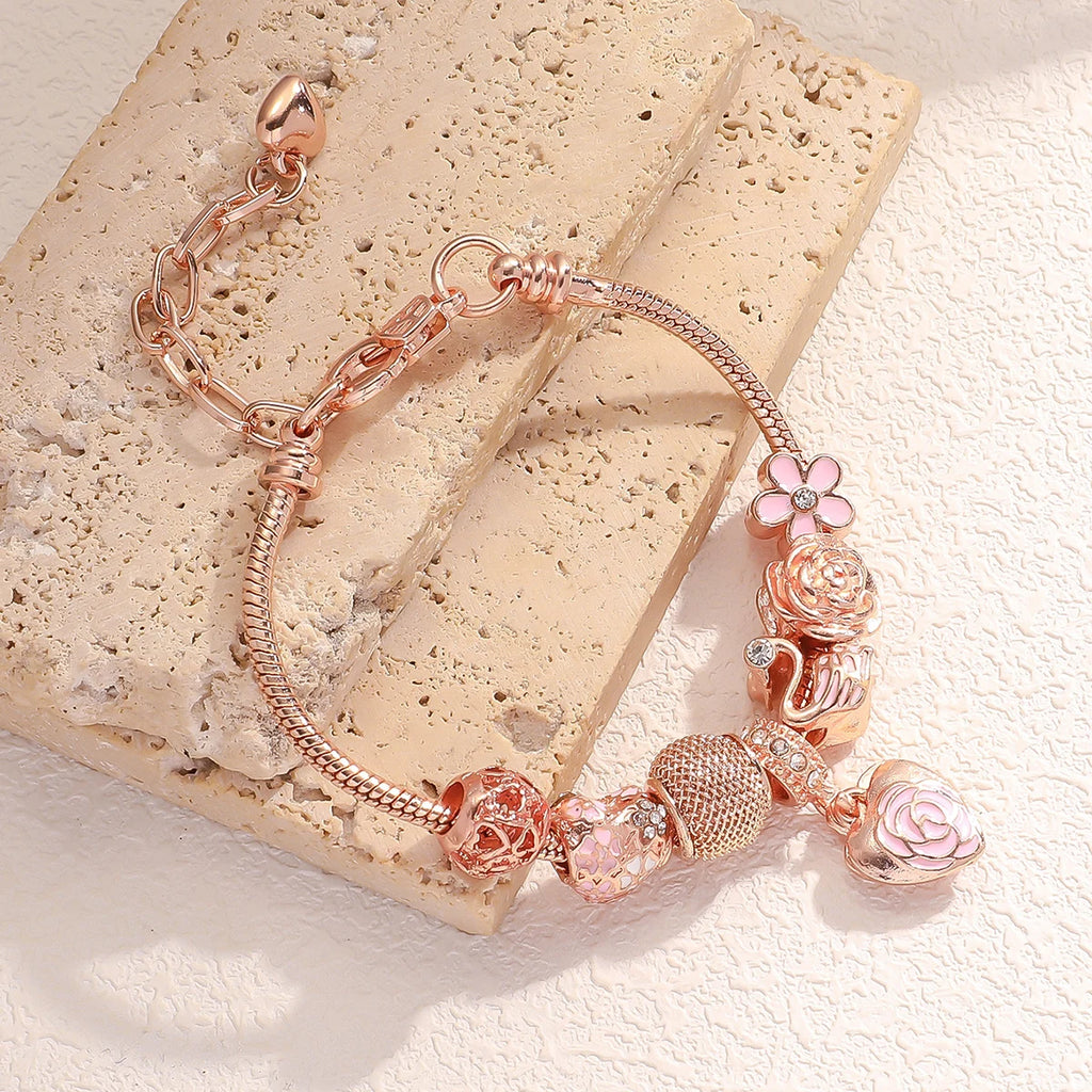 Rosette Swan Charm Bracelet displayed on textured stone, featuring rose gold chain and multi-element charms with floral and swan motifs.