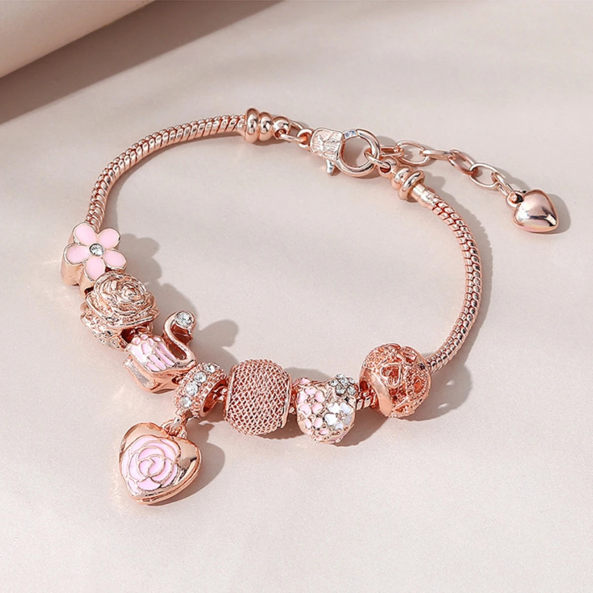 Rosette Swan Charm Bracelet in rose gold with pink enamel charms including a swan, rose, and heart, styled on a soft beige background.