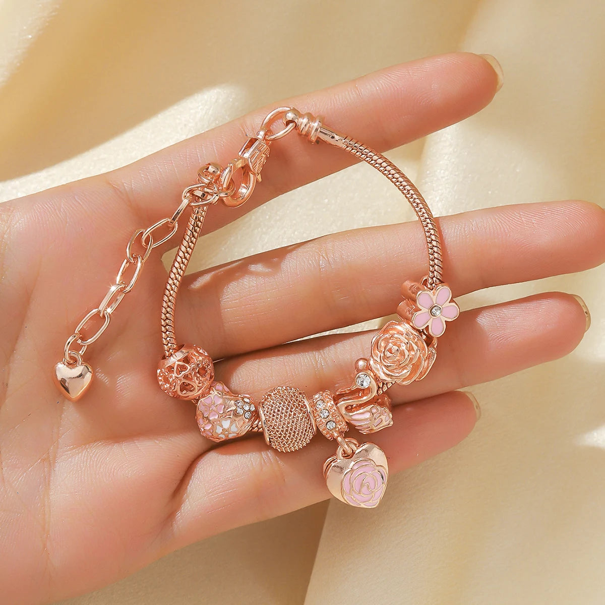 Rosette Swan Charm Bracelet held in hand, showcasing rose gold finish and detailed charms like a swan, flower, and heart with pink accents.